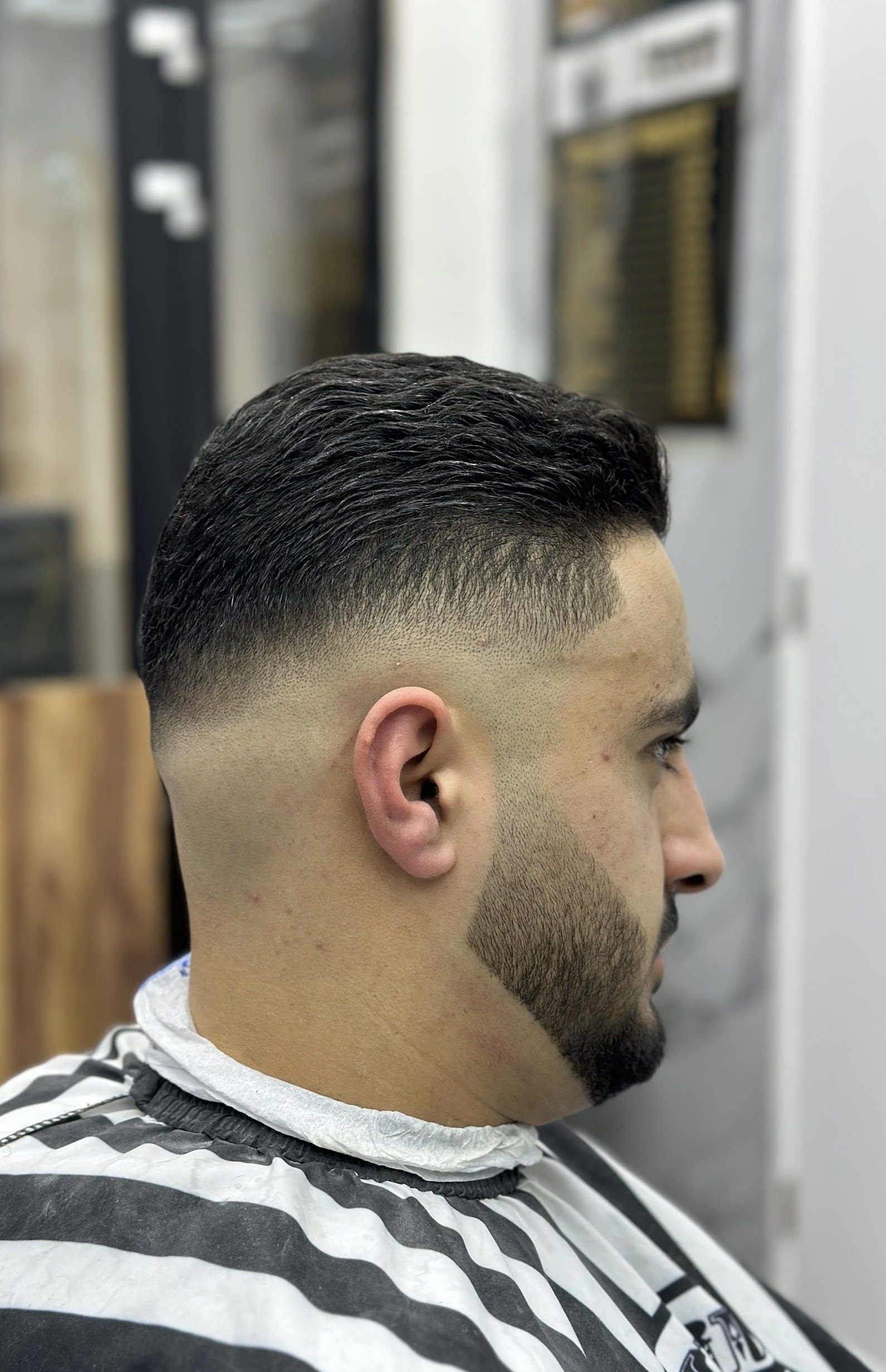 Zijaanzicht van een klant met een skin fade en een strak bijgewerkte baard, in een gestreepte kapmantel