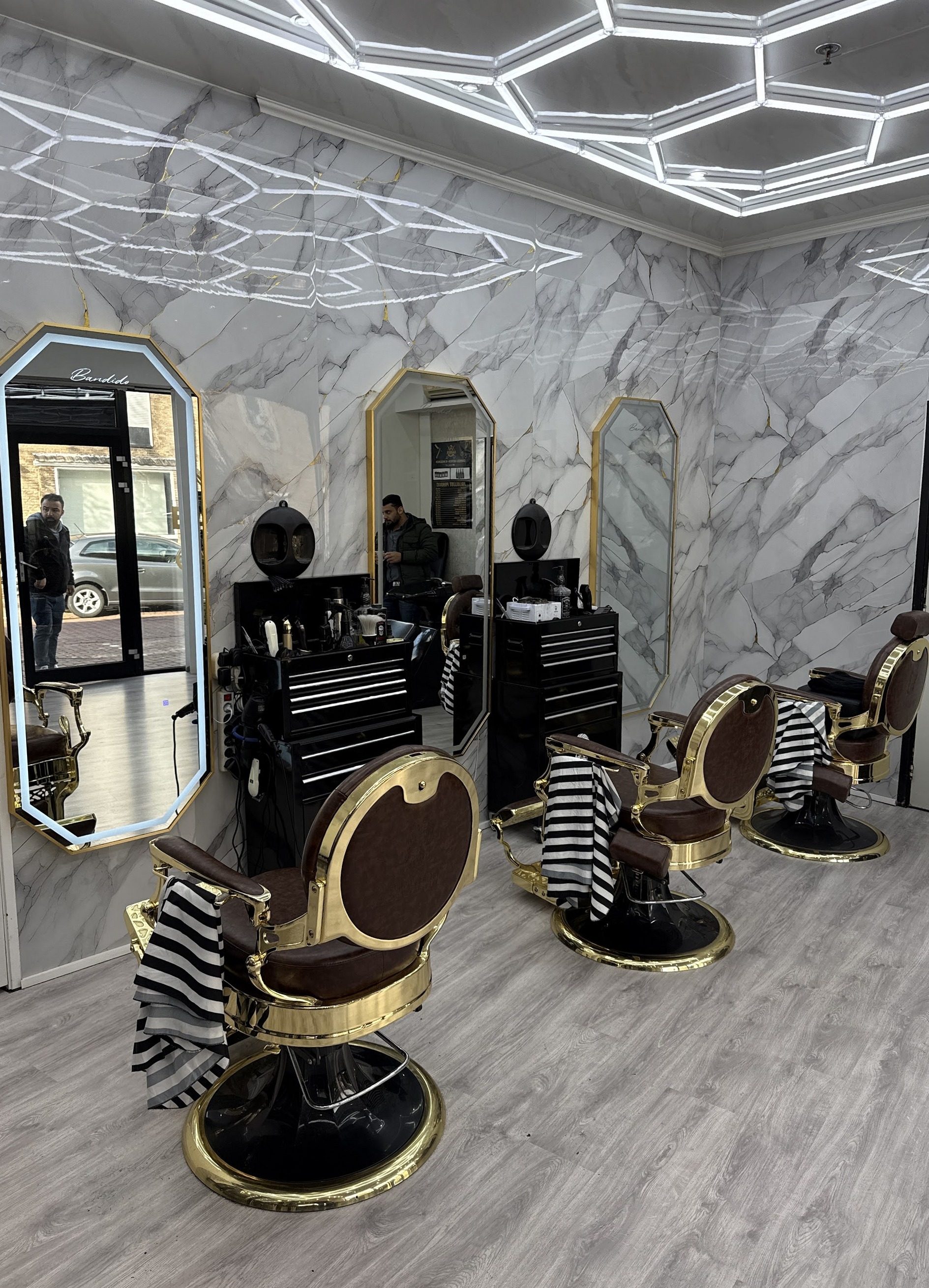 Het interieur van Barbershop Roosendaal: wit marmer op de wanden, goudomlijste achthoekige spiegels, een lichtplafond van zeshoeken en bruin met gouden barberstoelen