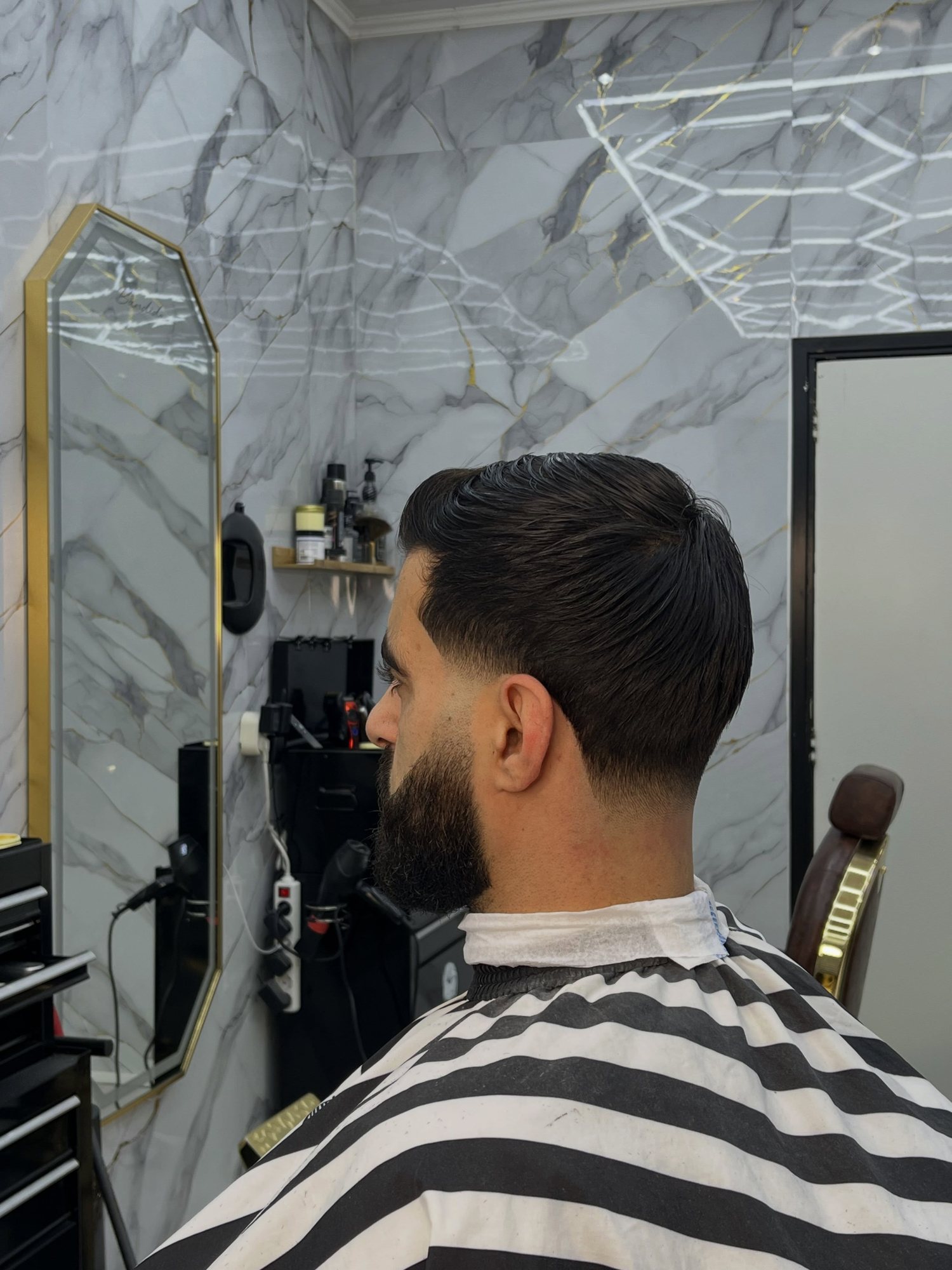 Klant van achteren gezien met een low fade en volle baard, voor de marmeren wand en een goudomlijste spiegel